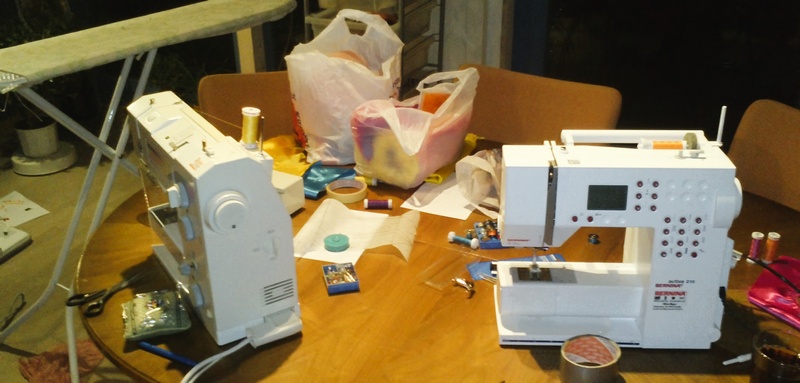 Zwei Nähmaschinen stehen auf dem Tisch. Dahinter ein Bügelbrett.